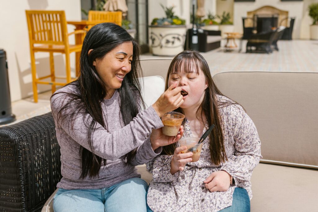 A mother joyfully feeds her daughter with Down syndrome on a sunny patio, sharing a snack together.
