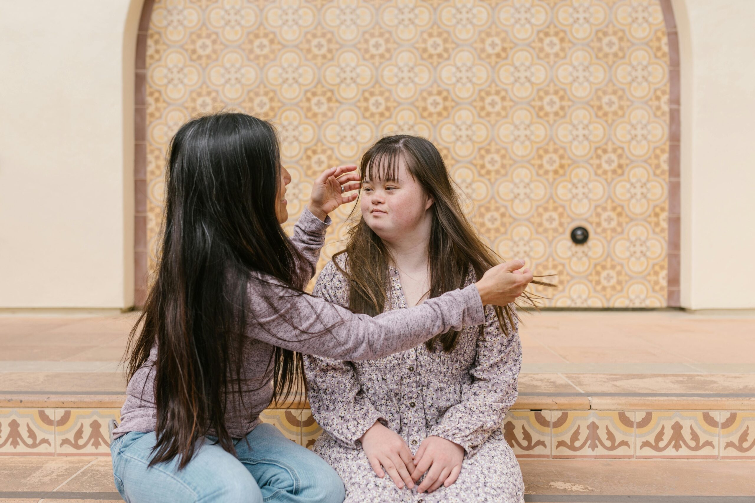 A mother lovingly adjusts her daughter's hair as they sit together on steps.